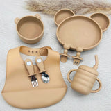 Vaisselle bébé beige en silicone alimentaire avec assiette, tasse, bol, bavoir et couverts adaptés.
